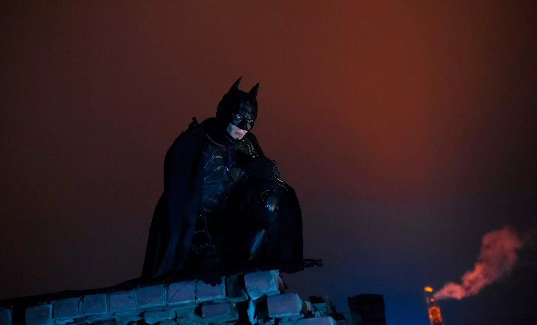 A man dressed as a bat in mask, helmet, armor and black cloak sits at the top of the building against the background of night lights and sky. Cosplay.