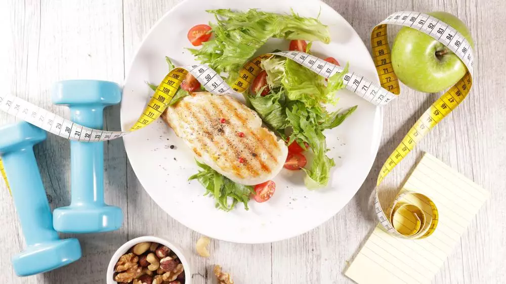 A healthy diet plate with grilled chicken and salad for weight loss with two dumbbells and the measuring tape kept alongside.
