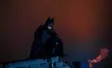 A man dressed as a bat in mask, helmet, armor and black cloak sits at the top of the building against the background of night lights and sky. Cosplay.