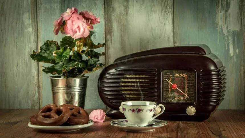 A vintage radio on a table with a flower vase, cookies and a tea cup.